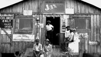 Movie still from “13th” (2016), directed by Ava DuVernay – A black and white photo of men sitting in front of a building; Extreme Wide shot, Low angle
