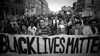 Movie still from “13th” (2016), directed by Ava DuVernay – A black lives matter banner is held up in front of a crowd of people; Wide shot, High angle