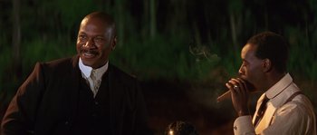 Movie still from “Rosewood” (1997), directed by John Singleton – A man in a black suit smoking a cigarette; Medium shot, Low angle