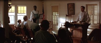 Movie still from “Rosewood” (1997), directed by John Singleton – A man standing in front of a group of people in a room; Medium shot, Over the shoulder angle