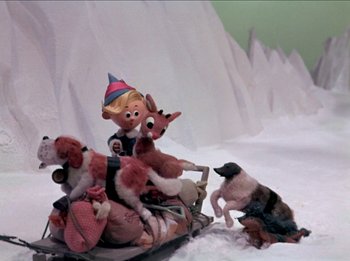 Movie still from “Rudolph the Red-Nosed Reindeer” (1964), directed by Larry Roemer – A stuffed animal bear is riding a sled with a dog; Wide shot, High angle