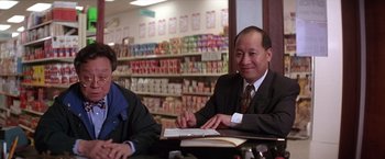 Movie still from “Rumble in the Bronx” (1995), directed by Stanley Tong – A man sitting at a table in front of shelves of food; Medium shot, Low angle