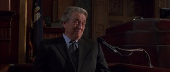 Movie still from “Runaway Jury” (2003), directed by Gary Fleder – An older man wearing a suit and tie while standing in front of a microphone; Close Up shot, Low angle