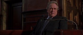 Movie still from “Runaway Jury” (2003), directed by Gary Fleder – A man in a suit and tie sitting in front of a microphone; Medium shot, Low angle