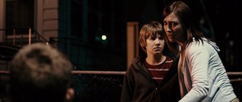 Movie still from “Running Scared” (2006), directed by Wayne Kramer – A woman standing next to a young boy in front of a chain link fence at night; Medium shot, Over the shoulder angle