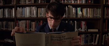 Movie still from “Rushmore” (1998), directed by Wes Anderson – A man in a suit and glasses reading a book in front of a bookcase; Close Up shot, High angle