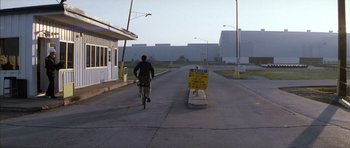 Movie still from “Rushmore” (1998), directed by Wes Anderson – A man riding a bike down a street next to buildings; Extreme Wide shot, Over the shoulder angle