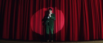 Movie still from “Rushmore” (1998), directed by Wes Anderson – A man standing on a stage in front of a red curtain; Wide shot, Overhead angle