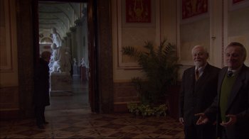 Movie still from “Russian Ark” (2002), directed by Aleksandr Sokurov – An older man standing in a hallway next to a potted plant; Wide shot, Over the shoulder angle
