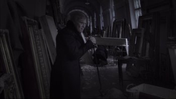 Movie still from “Russian Ark” (2002), directed by Aleksandr Sokurov – An older woman in a black coat is in a dark room; Medium shot, High angle