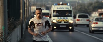 Movie still from “Rust and Bone” (2012), directed by Jacques Audiard – A man running on the side of the road while a ambulance is behind him; Medium shot, Over the shoulder angle