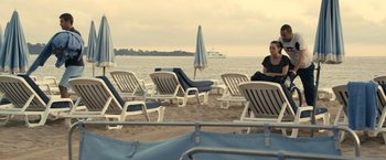 Movie still from “Rust and Bone” (2012), directed by Jacques Audiard – A person sitting on a beach next to a group of lounge chairs; Extreme Wide shot, High angle