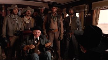 Movie still from “Rustlers' Rhapsody” (1985), directed by Hugh Wilson – A group of men standing in a room with a man sitting in a chair; Medium shot, Over the shoulder angle