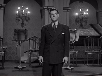 Movie still from “Saboteur” (1942), directed by Alfred Hitchcock – A man in a suit standing in front of chairs and chandeliers; Medium shot, Low angle