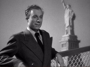 Movie still from “Saboteur” (1942), directed by Alfred Hitchcock – A black and white photo of a man in a suit and tie in front of the statue of liberty; Medium shot, Low angle