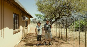 Movie still from “Safari” (2016), directed by Ulrich Seidl – A man and a woman posing for a picture in front of a building; Wide shot, Low angle