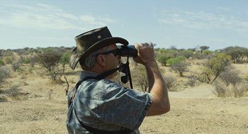 Movie still from “Safari” (2016), directed by Ulrich Seidl – A man with a hat on looking through binoculars in the desert; Medium shot, Over the shoulder angle