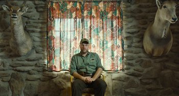 Movie still from “Safari” (2016), directed by Ulrich Seidl – A man sitting in a chair in front of a window; Medium shot, Low angle