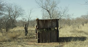 Movie still from “Safari” (2016), directed by Ulrich Seidl – A man standing in front of a wooden structure; Wide shot, Low angle