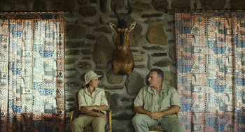 Movie still from “Safari” (2016), directed by Ulrich Seidl – A man and a woman sitting next to each other in front of an antelopes head mounted on the wall; Medium shot, Over the shoulder angle