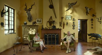 Movie still from “Safari” (2016), directed by Ulrich Seidl – A man and a woman sitting in chairs in a living room; Wide shot, High angle