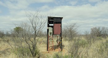 Movie still from “Safari” (2016), directed by Ulrich Seidl – A man standing in front of an outhouse in the middle of a field; Wide shot, Low angle