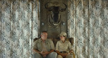 Movie still from “Safari” (2016), directed by Ulrich Seidl – A man and a woman sitting on a couch in front of an animal head; Wide shot, Over the shoulder angle