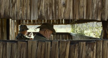 Movie still from “Safari” (2016), directed by Ulrich Seidl – An older man wearing a cowboy hat looking through a fence; Medium shot, Over the shoulder angle