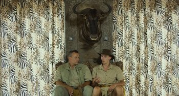 Movie still from “Safari” (2016), directed by Ulrich Seidl – Two men sitting in front of a wall with a buffalo on it; Medium shot, Over the shoulder angle