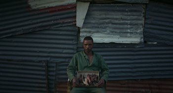 Movie still from “Safari” (2016), directed by Ulrich Seidl – A man holding an old picture in front of an old building; Medium shot, Low angle
