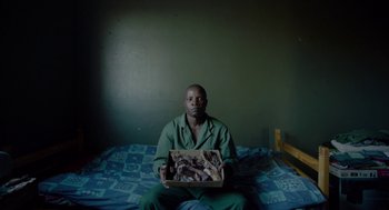 Movie still from “Safari” (2016), directed by Ulrich Seidl – A man sitting on top of a bed holding a box; Medium shot, Overhead angle