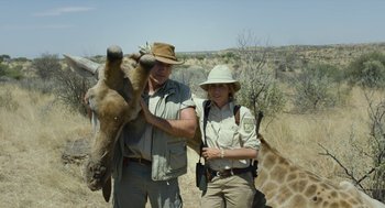 Movie still from “Safari” (2016), directed by Ulrich Seidl – A man and a woman standing next to a giraffe; Medium shot, Low angle
