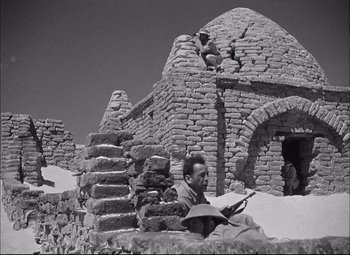 Movie still from “Sahara” (1943), directed by Zoltan Korda – A man sitting on a rock wall reading a book; Wide shot, Low angle