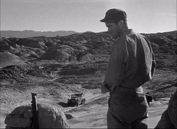 Movie still from “Sahara” (1943), directed by Zoltan Korda – A man standing on a dirt hill looking at an army vehicle; Wide shot, Over the shoulder angle