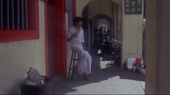 Movie still from “Saint Jack” (1979), directed by Peter Bogdanovich – A man sitting on a stool in front of a building; Wide shot, High angle