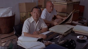 Movie still from “Saint Jack” (1979), directed by Peter Bogdanovich – Two men sitting at a table with a computer; Medium shot, High angle