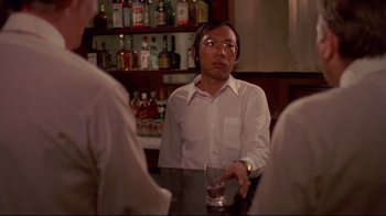 Movie still from “Saint Jack” (1979), directed by Peter Bogdanovich – A man sitting at a table in front of two other men; Medium shot, Over the shoulder angle