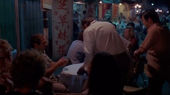 Movie still from “Saint Jack” (1979), directed by Peter Bogdanovich – A group of people sitting around a table with food on it; Wide shot, Over the shoulder angle