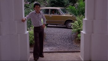 Movie still from “Saint Jack” (1979), directed by Peter Bogdanovich – A man standing in front of a driveway with a car in the background; Wide shot, Low angle