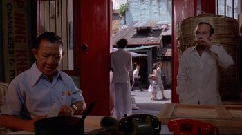 Movie still from “Saint Jack” (1979), directed by Peter Bogdanovich – A man sitting at a table in front of a red door; Medium shot, Over the shoulder angle