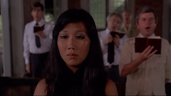 Movie still from “Saint Jack” (1979), directed by Peter Bogdanovich – A woman with long black hair is looking at the camera while a group of people are taking pictures in the background; Close Up shot, Over the shoulder angle