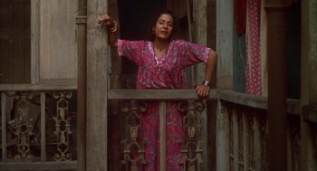 Movie still from “Salaam Bombay!” (1988), directed by Mira Nair – A woman in a pink dress standing next to an iron gate; Medium shot, Low angle