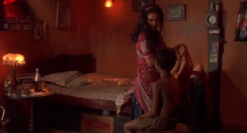 Movie still from “Salaam Bombay!” (1988), directed by Mira Nair – A young boy sitting on a bed next to a woman; Wide shot, High angle