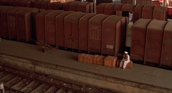 Movie still from “Salaam Bombay!” (1988), directed by Mira Nair – A man sitting on top of a crate next to a bunch of train cars; Extreme Wide shot, High angle