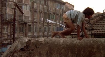 Movie still from “Salaam Bombay!” (1988), directed by Mira Nair – A man with a baseball bat in his hand is standing on the side of a building; Wide shot, Low angle