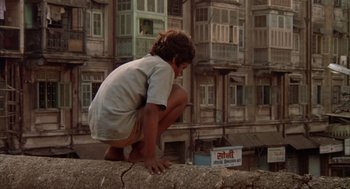 Movie still from “Salaam Bombay!” (1988), directed by Mira Nair – A young boy squatting on the side of a building; Wide shot, Overhead angle