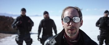 Movie still from “Salt and Fire” (2016), directed by Werner Herzog – A man wearing sunglasses while standing next to two other men; Close Up shot, Over the shoulder angle