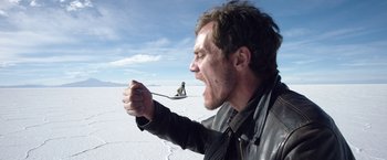 Movie still from “Salt and Fire” (2016), directed by Werner Herzog – A man is holding a spoon in his mouth while sitting on the ground; Medium shot, Low angle