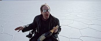 Movie still from “Salt and Fire” (2016), directed by Werner Herzog – A man sitting on the ground holding a bottle of wine; Medium shot, High angle