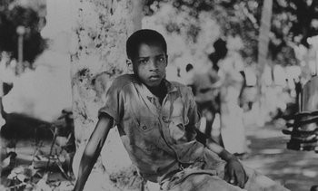 Movie still from “Salut les Cubains” (1963), directed by Agnès Varda – A black and white photo of a young boy sitting in front of a tree; Medium shot, High angle